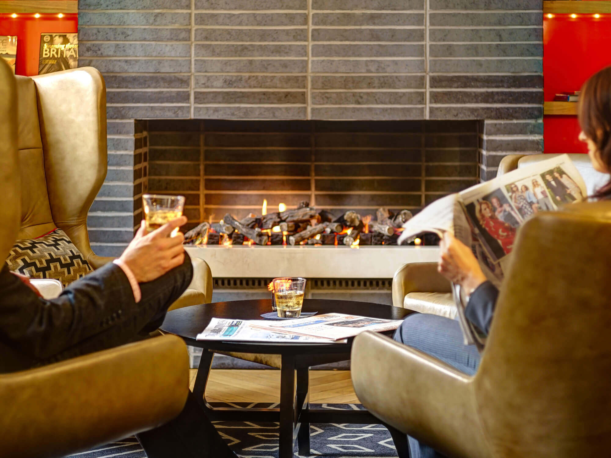 Two people drinking and reading the newspaper by the fireplace